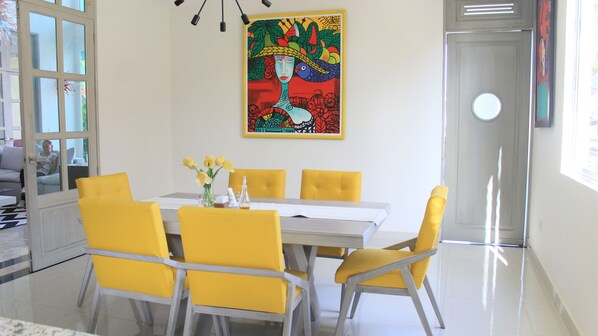 Un coin repas avec une table blanche et des chaises jaunes, un tableau coloré au mur et une porte sur la droite.