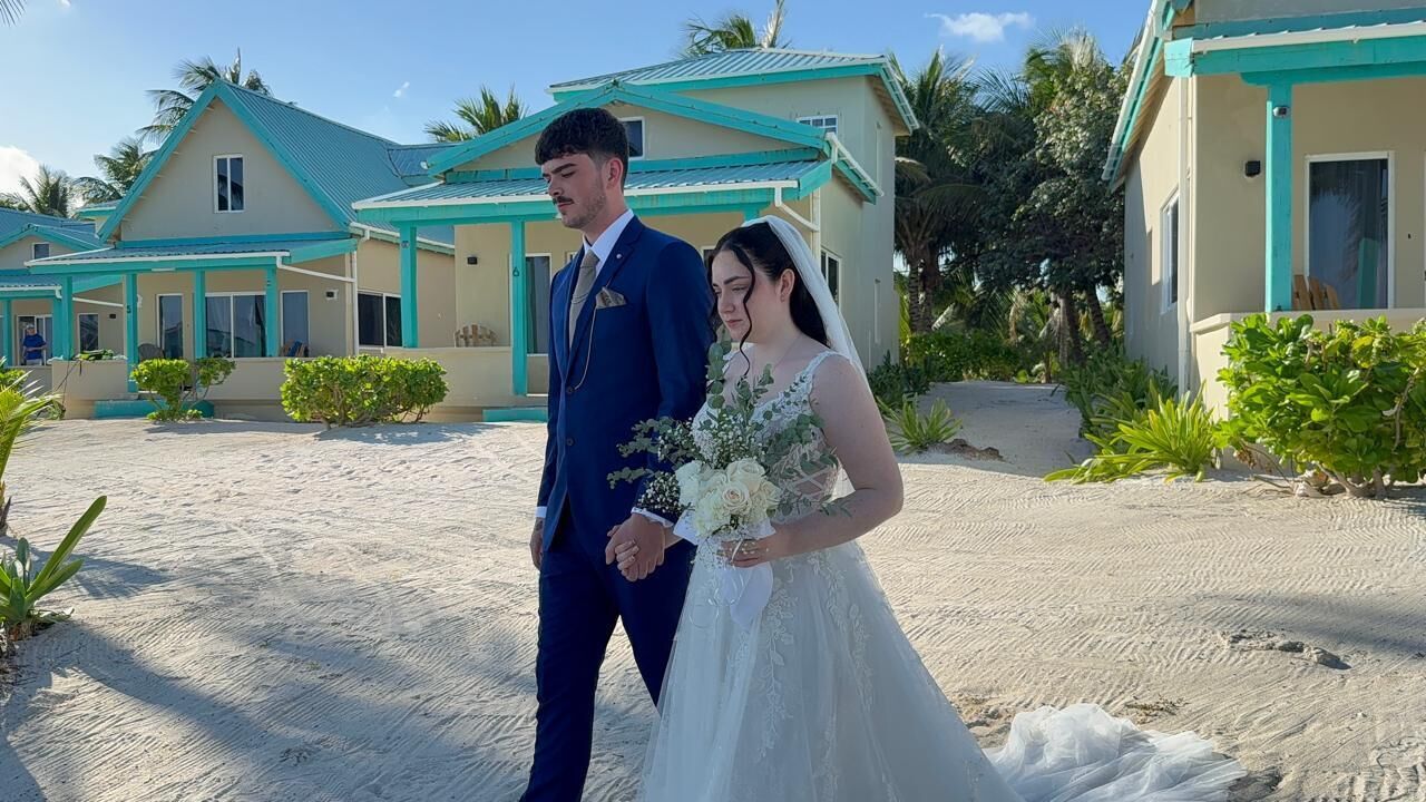 A couple standing in front of a resort with a sandy beach and turquoise buildings.