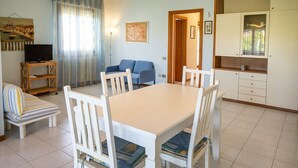 A dining area with a table and chairs, a sofa, a television on a stand, and a kitchen area with cabinets. - S220 - Sirolo, three-room apartment in the center with terrace (Sirolo)