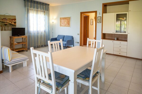 A dining area with a table and chairs, a sofa, a television on a stand, and a kitchen area with cabinets.