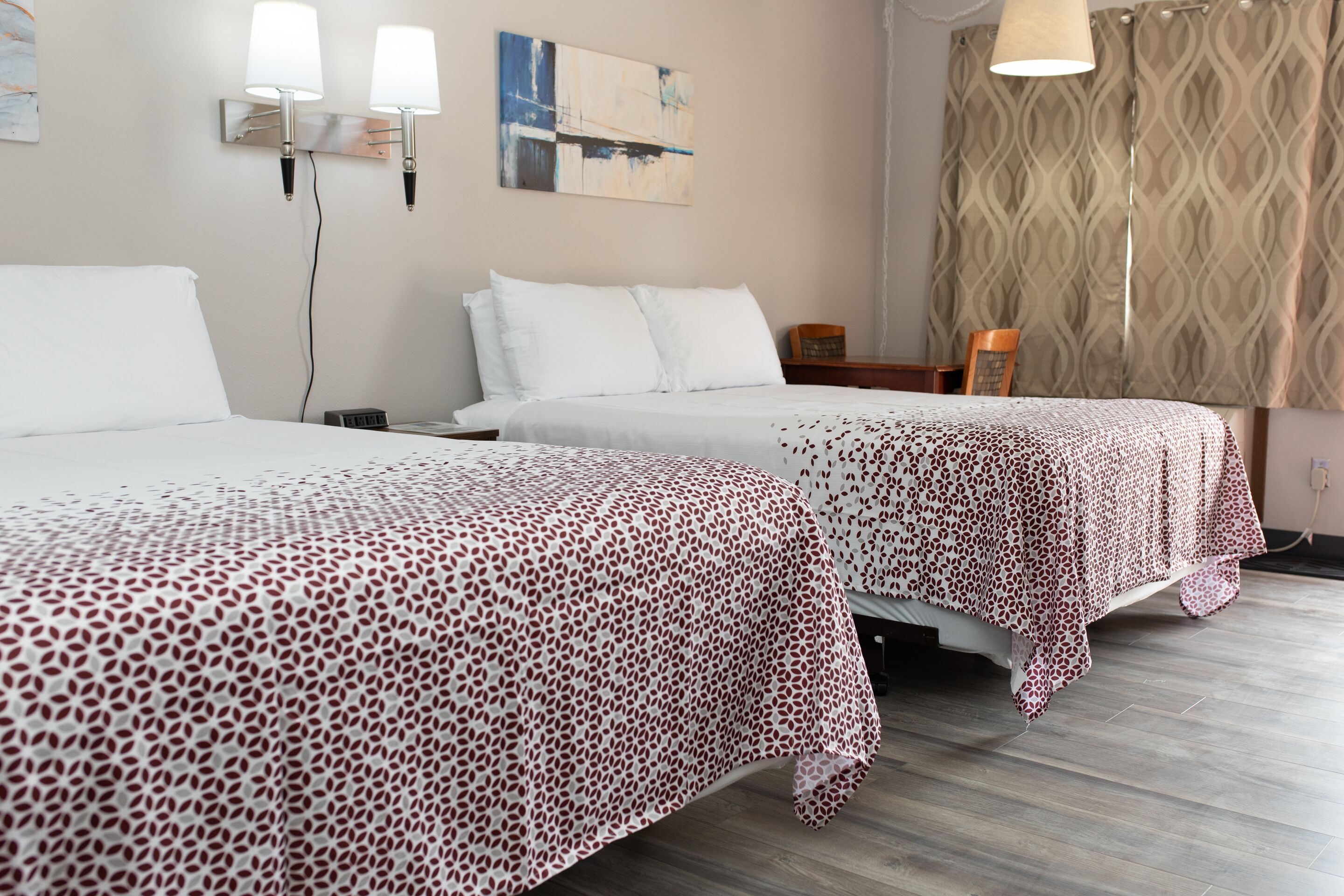 A hotel room with two beds, a patterned bedspread, a wall-mounted lamp, and a patterned curtain.