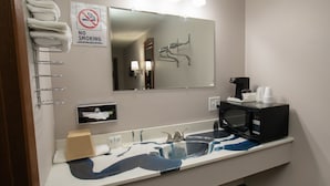 Un bany amb un lavabo, un mirall, un suport de tovalloles, senyalització de prohibició de fumar i un microones.