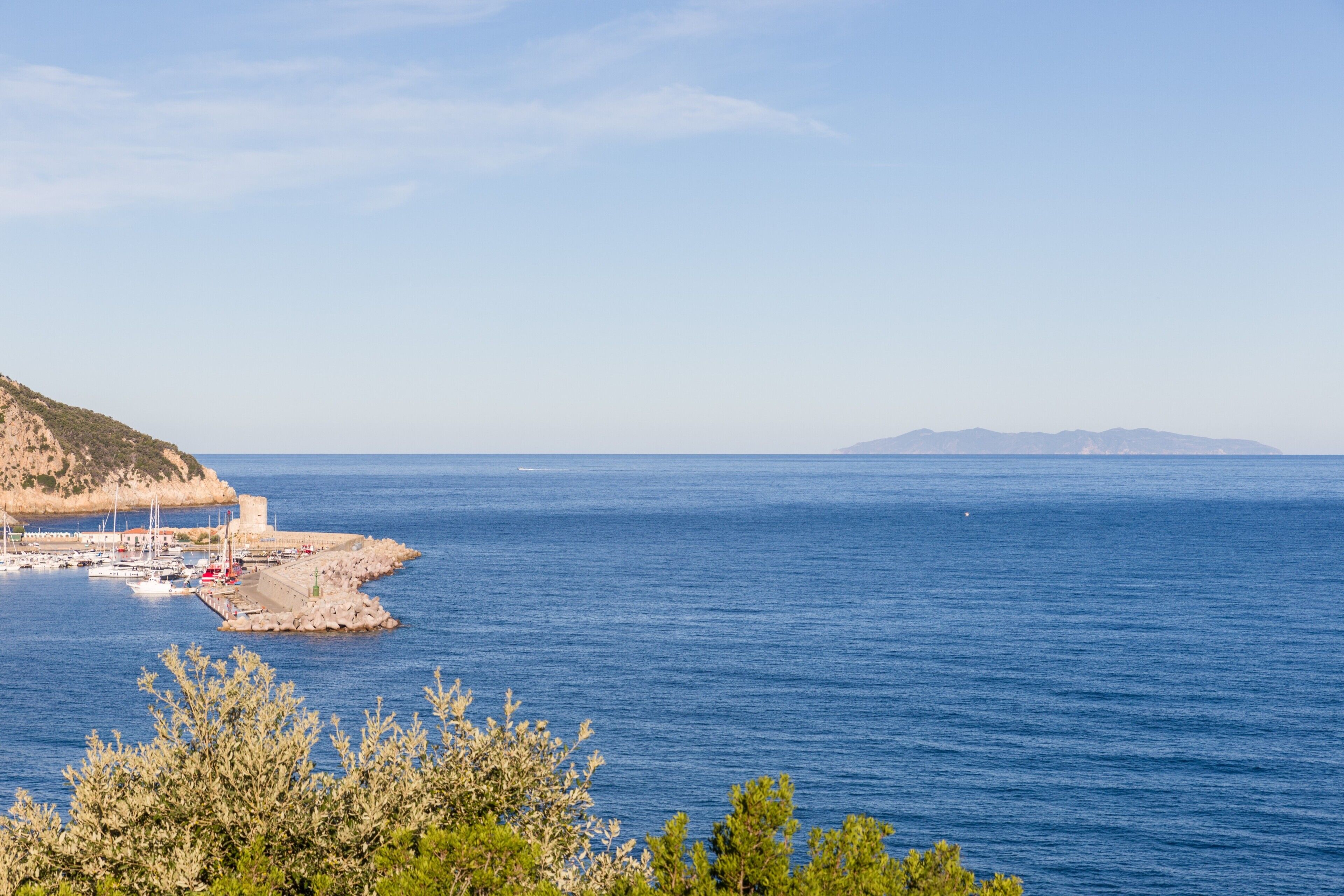 Una marina costera con barcos, un litoral rocoso y montañas a la distancia.