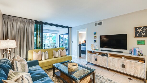 A modern living room with a sofa, a coffee table, a TV, and a large window. - Beachy Marco Island Condo with Resort Amenities! (Marco Island)
