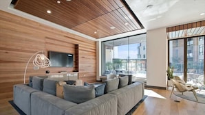 Ein modernes Wohnzimmer mit einem großen, grauen Ecksofa, einem flachbildfernseher, der an einer Holzwand angebracht ist, und einem großen Fenster mit Blick auf die Stadt.