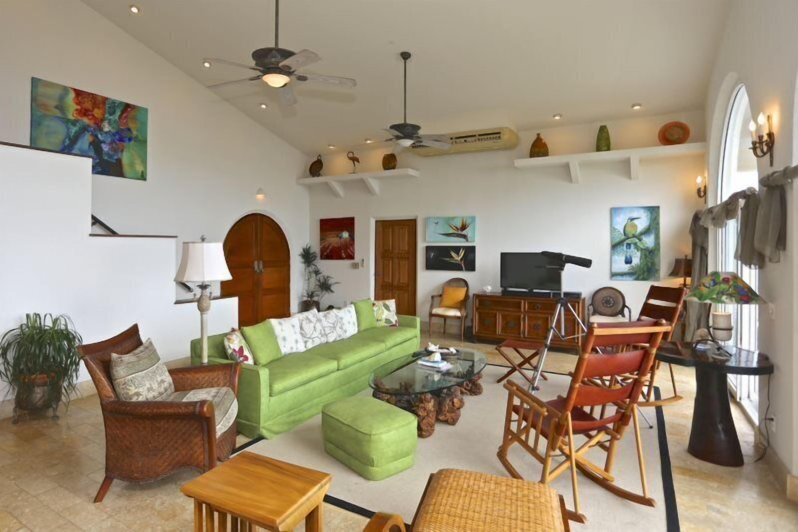 A spacious living room with a green sofa, wooden chairs, a coffee table, and a TV. There are decorative items on shelves and a ceiling fan.