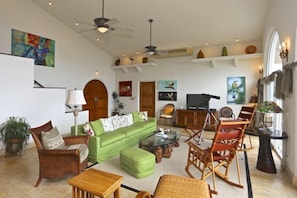 Un salon spacieux avec un canapé vert, des chaises en bois, une table basse et un téléviseur. Des objets décoratifs sont disposés sur des étagères et un ventilateur de plafond est installé.