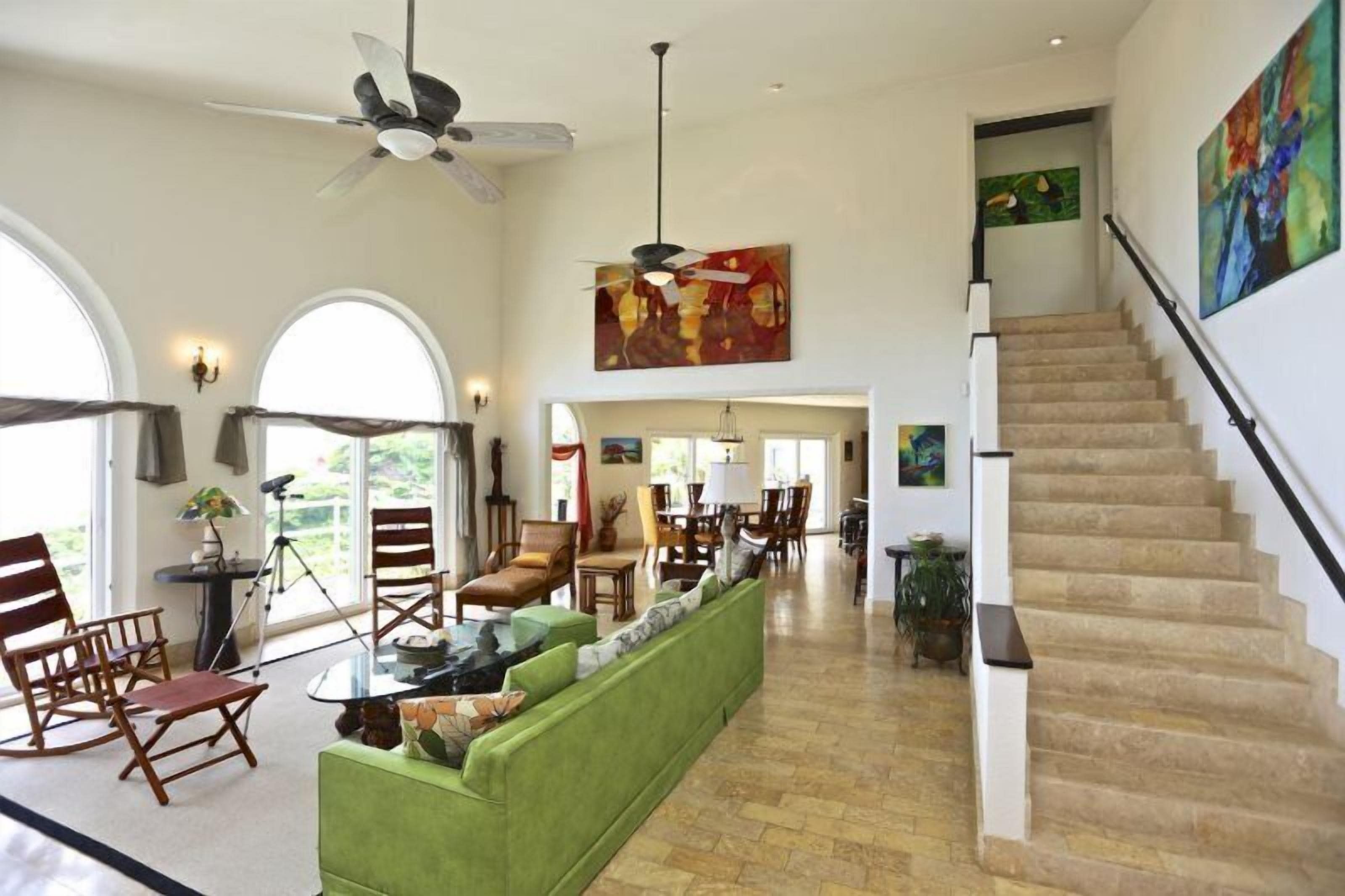 A spacious living room with a green sofa, wooden chairs, a dining area, and a staircase.