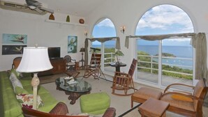 A spacious living room with a large window offering a view of the sea. There is a green sofa, a coffee table, and several chairs. - Villa Cielo Azul (Sardinal)