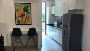 A modern hotel room with a bed, a desk, two chairs, a refrigerator, and a kitchen area with cabinets and a microwave.