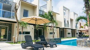 A modern poolside area with lounge chairs and a large umbrella, surrounded by palm trees and a pool.