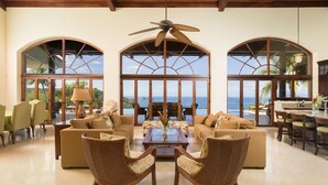 A spacious living room with a large arched window, a ceiling fan, and a view of the ocean. - Cinco Sinsotes (Sardinal)