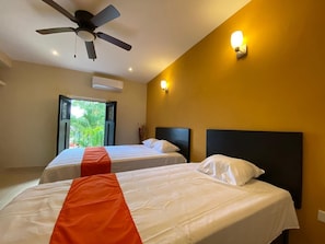 Habitación de hotel con dos camas, ventilador de techo y ventana con vistas a zonas verdes.