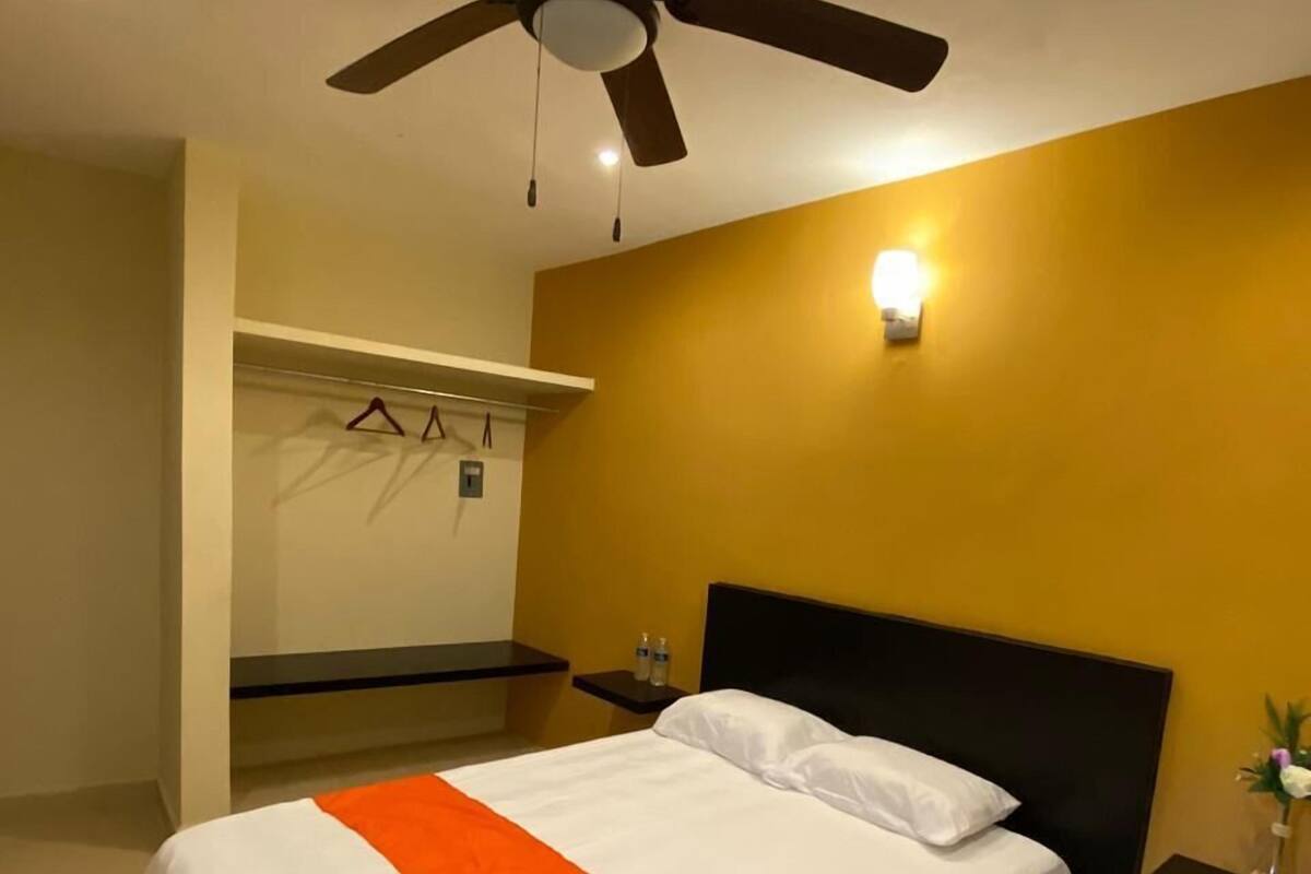A hotel room with a bed, a ceiling fan, a nightstand, a wall-mounted light, and a mirror.