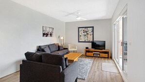 Un salon moderne avec un canapé gris, une table basse en bois, un téléviseur posé sur un meuble en bois et des œuvres d’art accrochées aux murs.