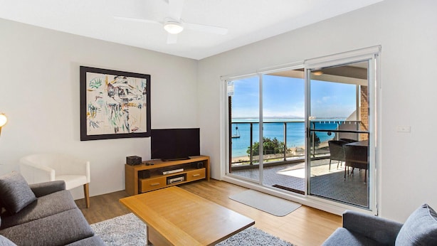 Un salon moderne avec un téléviseur à écran plat, une table basse en bois et un canapé. Une grande fenêtre offre une vue sur la mer, et un balcon est aménagé avec du mobilier d’extérieur.