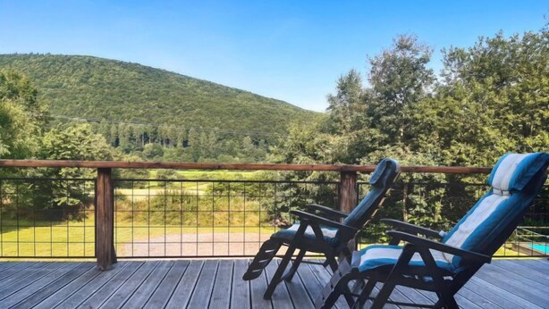 Une terrasse en bois avec deux chaises longues et une table, offrant une vue sur un paysage verdoyant et une colline.