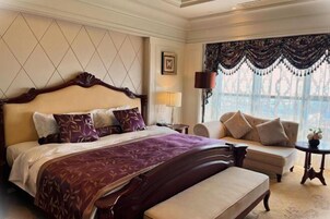 Una habitación de hotel con una cama grande, un sofá, un banco, una mesita de noche con lámpara y una ventana con cortinas.