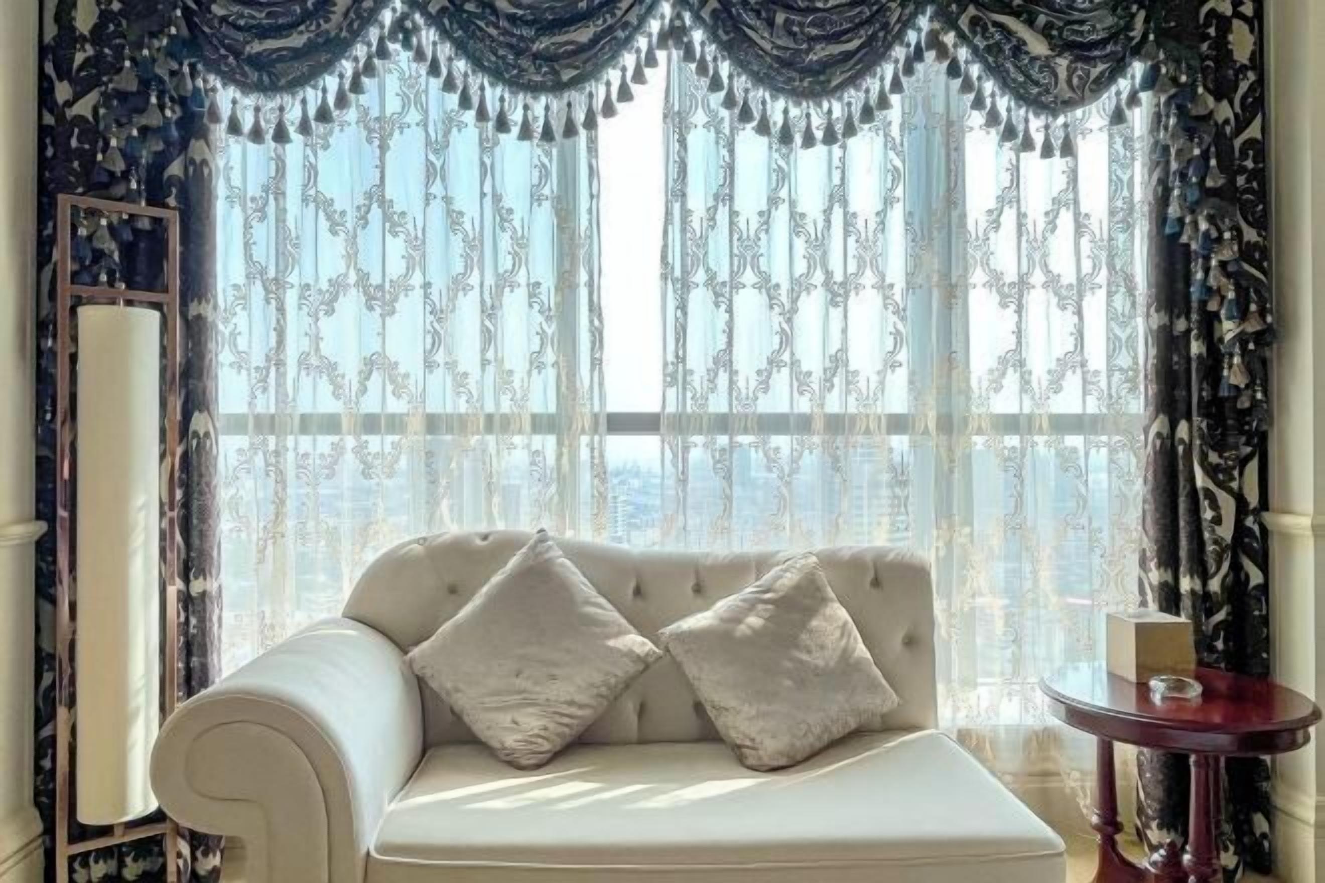 Une pièce avec un canapé blanc, deux coussins décoratifs, une table ronde avec une bougie et une grande fenêtre ornée de rideaux à motifs.