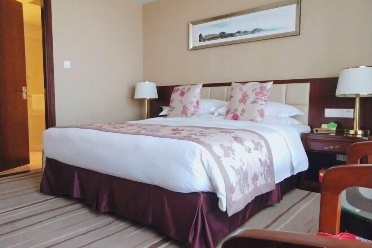 Habitación de hotel con una cama grande, dos mesitas de noche, una silla, un cuadro en la pared y piso alfombrado.