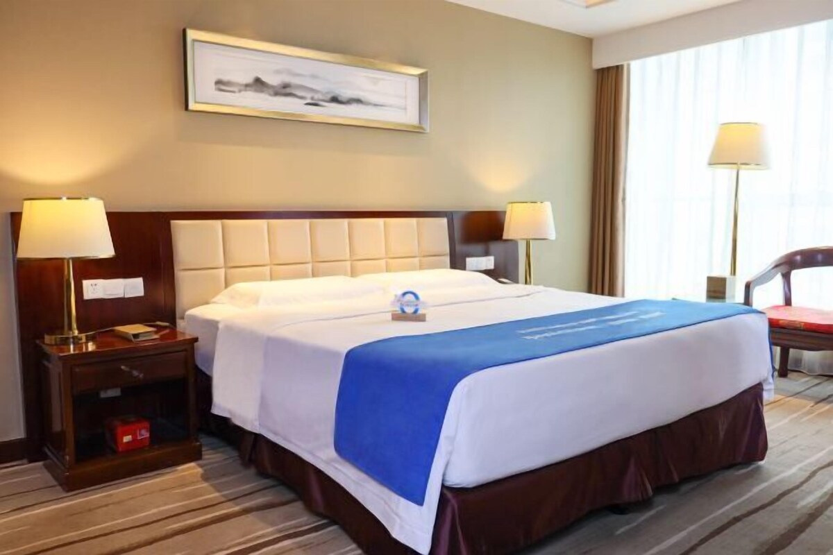 Habitación de hotel con una cama grande, dos mesitas de noche con lámparas, un cuadro enmarcado en la pared y una ventana con cortinas.