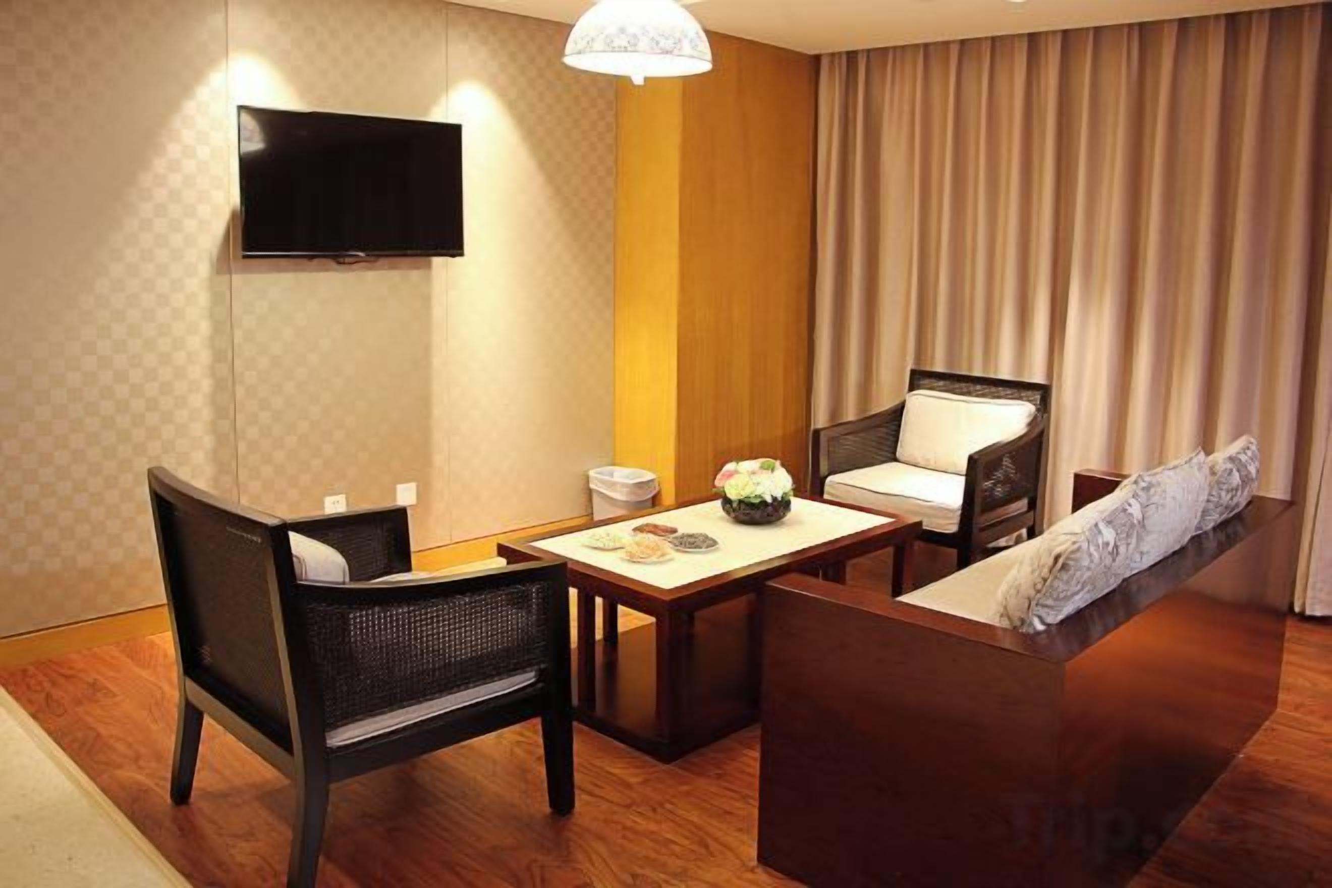 A hotel room with a TV mounted on the wall, a dining table with chairs, a sofa, and a large window with curtains.