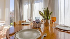 Un coin repas avec une table en bois dressée pour deux personnes, une plante en pot et une vue sur la ville à travers de fines rideaux.