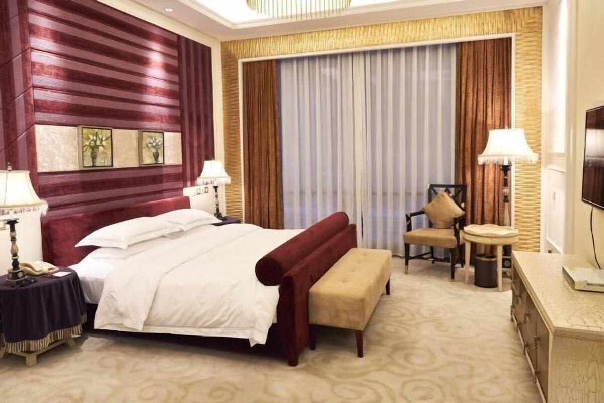 A hotel room with a large bed, a bedside table with a lamp, a chair, a small round table, and a TV.