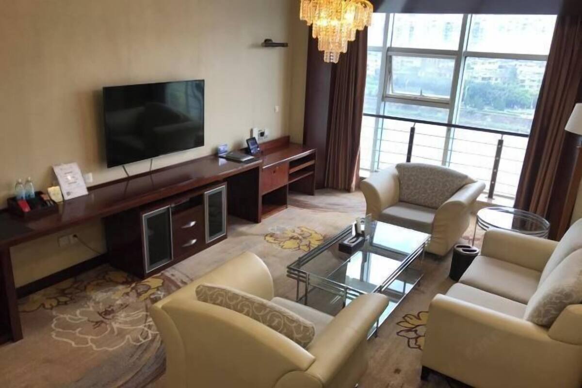 A living room with a flat-screen TV, a glass coffee table, two beige sofas, a wooden entertainment unit, and a large window with curtains.