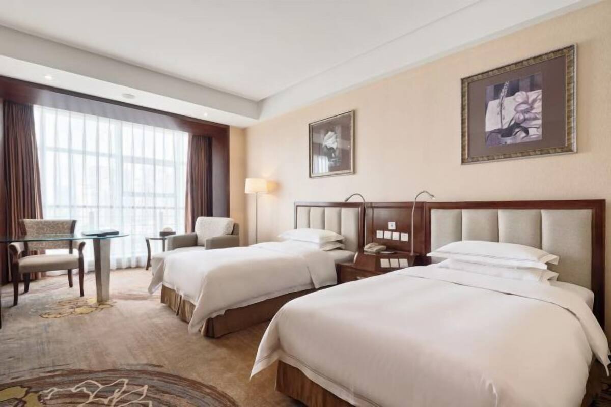 A hotel room with two beds, a desk, a chair, and a large window with curtains.
