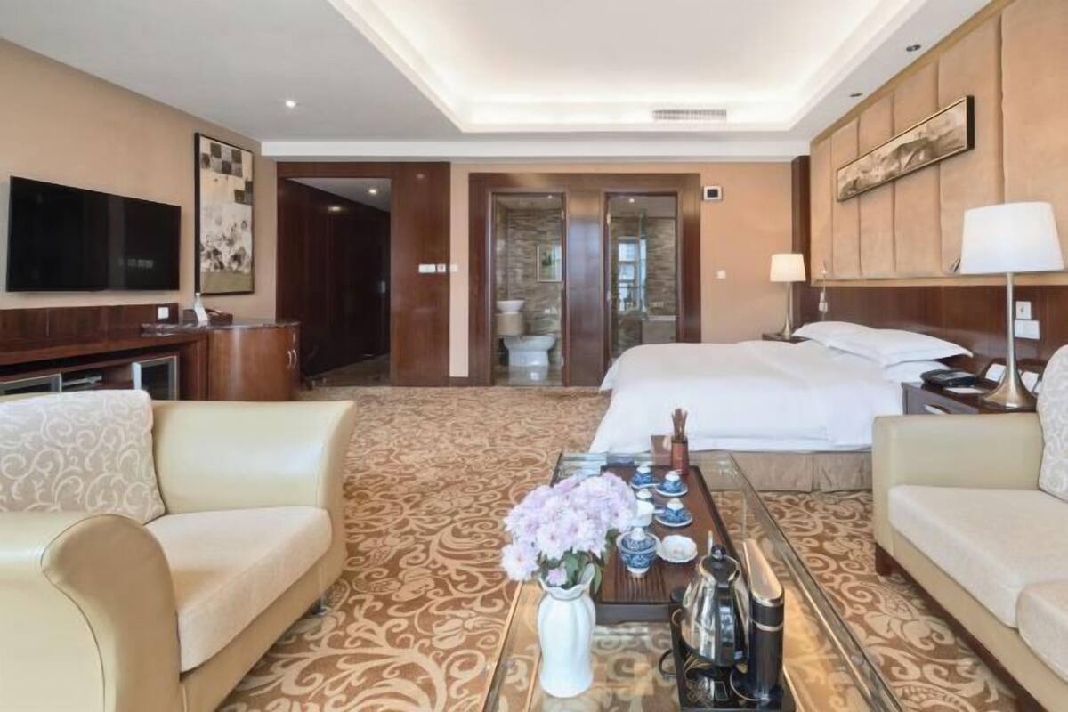 A hotel room with a large bed, two sofas, a coffee table with flowers, a TV, and a bathroom visible through a glass door.