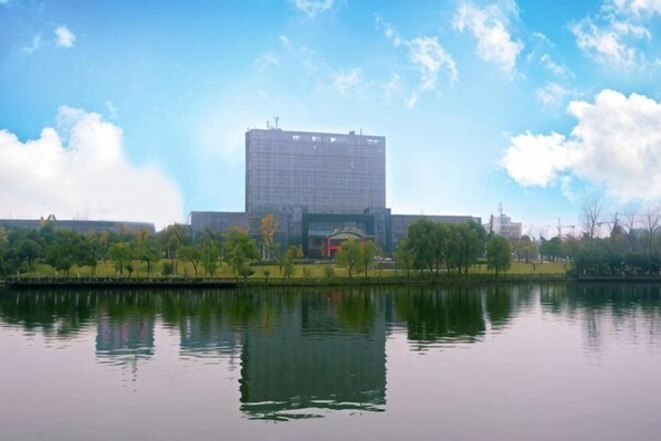 Un edificio grande reflejado en aguas tranquilas, rodeado de árboles y un cielo despejado.