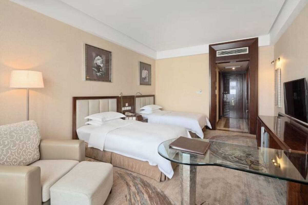 A hotel room with two beds, a sofa, a glass coffee table, a TV, and a hallway leading to another room.