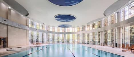 An indoor swimming pool with clear glass walls, a central diving platform, and a ceiling with a sky-like design.