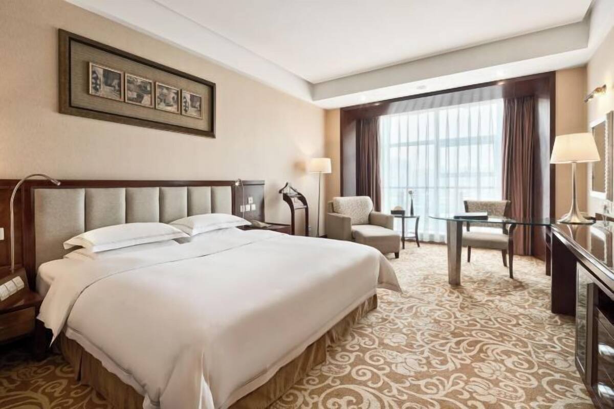 A hotel room with a large bed, a desk, a chair, and a large window with curtains.