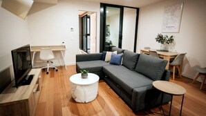 Un salon moderne avec un canapé d’angle gris, une petite table basse blanche, un téléviseur posé sur un meuble en bois, un coin repas avec une table et des chaises en bois, et une carte accrochée au mur.