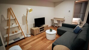 Un salon moderne avec un plancher en bois, un téléviseur à écran plat, un canapé gris foncé, une table basse ronde, un bureau avec une chaise et une lampe blanche.