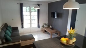 Een woonkamer met een bank, een salontafel en een televisie. Er is een raam met gordijnen, een plant en een fruitschaal op de tafel.