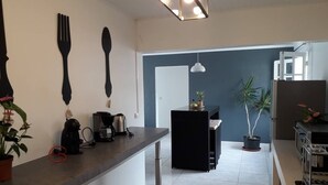 Een moderne keuken met een lange aanrecht, een koffieapparaat en een kleine koelkast. Er staan potplanten en er hangt een lamp.