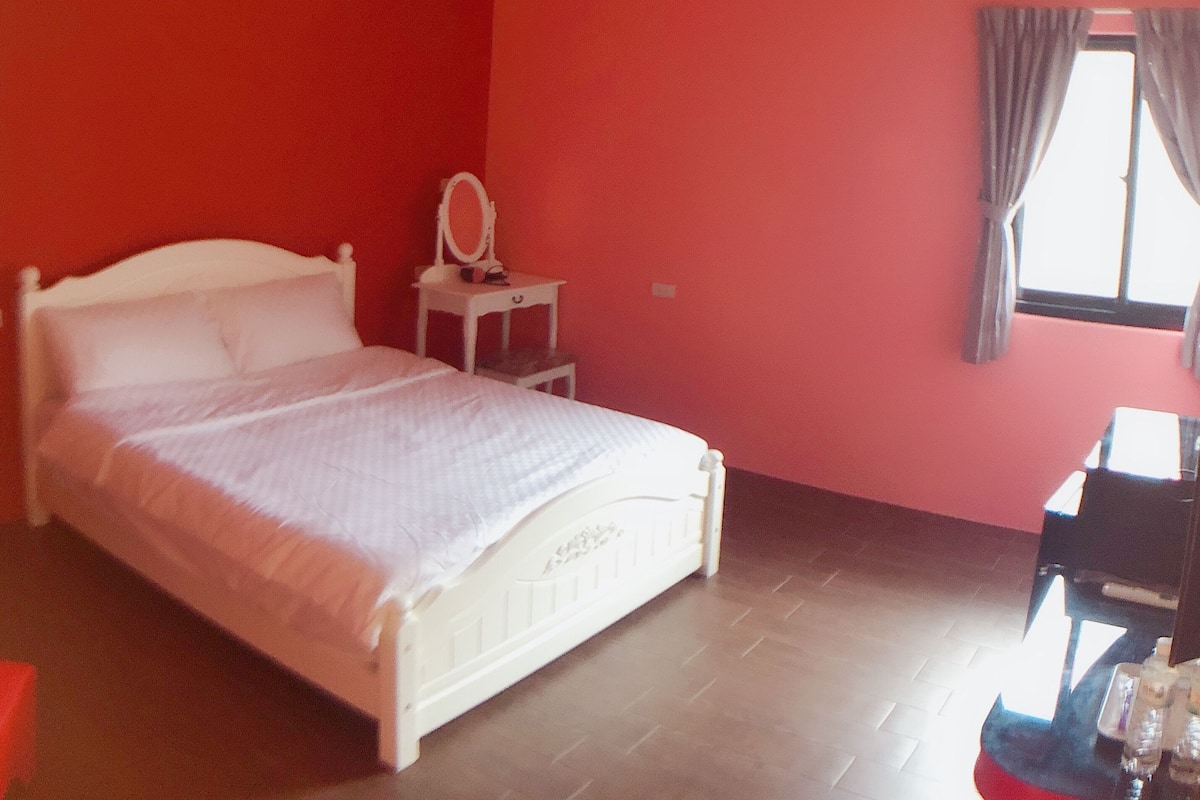 A bedroom with a white bed, a small bedside table with a mirror, and a window with curtains.
