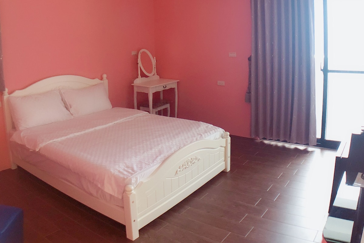 A bedroom with a bed, a small bedside table, a mirror, and a window with curtains.