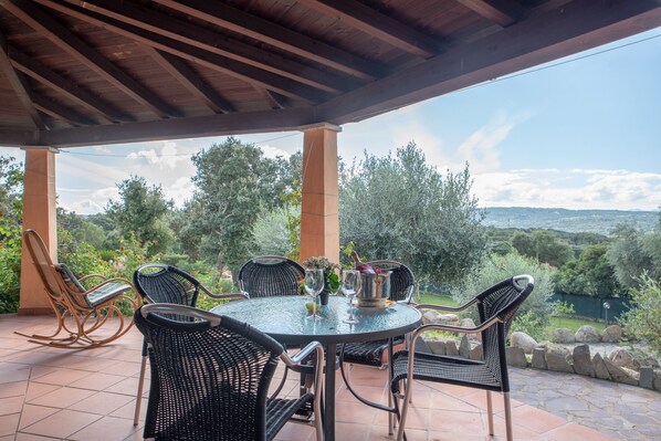 Une terrasse couverte avec une table ronde, des chaises en osier noir et une vue sur un lac et des collines.