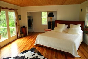 A bedroom with a wooden floor, a large bed with white bedding, a lamp, and a view of greenery outside.