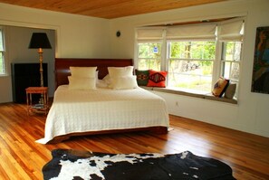 A bedroom with a wooden floor, a bed with white bedding, a window with a view of greenery, and a small table with a lamp.