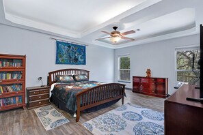 Un dormitorio con una cama, ventilador de techo, estantería, cómoda y un gran ventanal.