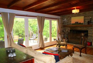 A cozy living room with a fireplace, comfortable seating, and large windows offering a view of the outdoors. - Romantic country cottage right on the Cypress Creek! (Wimberley)
