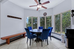 Un comedor con una mesa redonda, seis sillas azules, un banco de madera, un ventilador de techo y ventanas grandes con vistas a árboles.