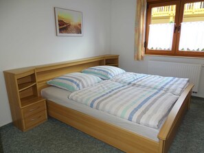 Habitación individual con una cama de una sola plaza, cabecera de madera, mesita de noche con cajones, radiador y ventana con cortinas.
