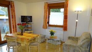 Ein Wohnzimmer mit Esstisch, Stühlen, einer Couch, einem Fernseher und einem Fenster mit Vorhängen.