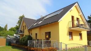 Ein gelbes Haus mit Balkon, Solaranlage und einem von einer Hecke umgebenen Garten mit Blumen.